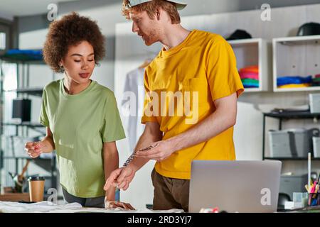 Jeune artisan tenant la couche d'impression et parlant à un collègue afro-américain tout en travaillant ensemble près de vêtements, café à emporter et ordinateur portable sur table Banque D'Images