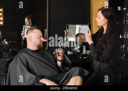 Le maître coiffeur montre au client une nouvelle coiffure dans le miroir. Homme avec un verre de boisson Banque D'Images