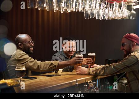 Barman barbu en lunettes de soleil, bonnet beanie et chemise passant deux verres de bière à des copains masculins interculturels joyeux debout près du comptoir du bar Banque D'Images