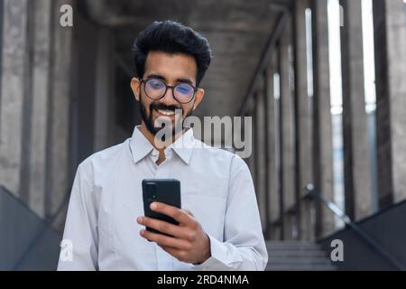 Jeune homme d'affaires hispanique marchant à l'extérieur de l'immeuble de bureaux dans la ville, homme dans la chemise tenant le téléphone, satisfait de résultat de réalisation freelancer, en utilisant l'application en ligne. Banque D'Images