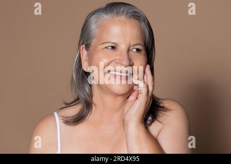 Souriante vieille dame caucasienne avec les cheveux gris touche le visage avec la main, apprécie le traitement de routine anti-âge Banque D'Images