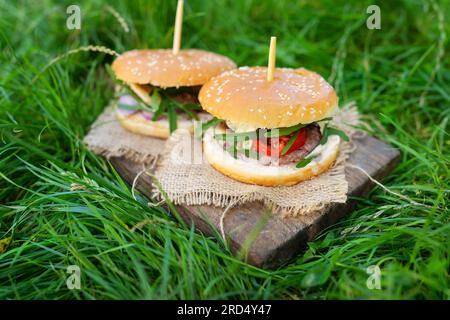 Délicieux hamburgers frais servis sur des planches de bois, photo à l'extérieur sur de l'herbe verte. Gros plan Banque D'Images