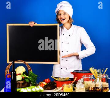 Chef féminin avec des légumes à la table tenant le tableau noir de menu blanc. Belle femme chef avec tableau noir vierge. Cuisinez en uniforme de chef à la cuisine Banque D'Images