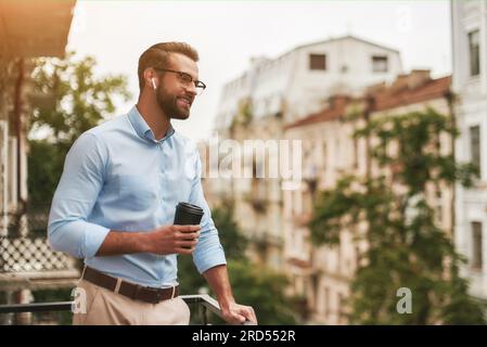Bonne nouvelle. Jeune et bel homme barbu dans des lunettes et des écouteurs tenant une tasse de café et parlant avec un ami tout en se tenant au bureau Banque D'Images