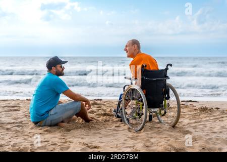 Portrait une personne handicapée en fauteuil roulant sur la plage avec un ami assis regardant la mer Banque D'Images