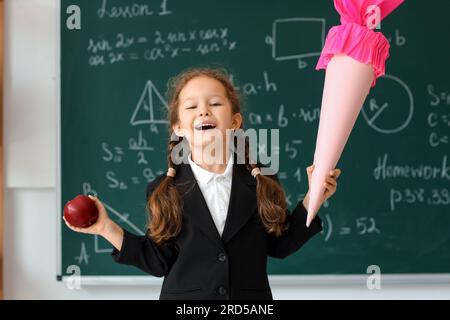 Petite fille heureuse avec cône d'école rose et pomme fraîche dans la salle de classe près du tableau noir Banque D'Images