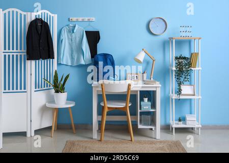 Intérieur du lieu de travail avec bureau, étagère, écran pliant, horloge et uniforme scolaire élégant accroché sur le mur bleu Banque D'Images