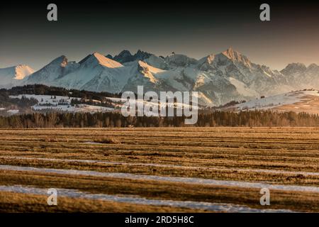 Montagnes rocheuses enneigées, terres agricoles et villages. ciel nuageux assombrissant, au coucher du soleil. Banque D'Images