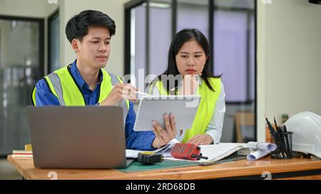 Deux ingénieurs asiatiques professionnels et sérieux portant des gilets de sécurité regardent l'écran d'une tablette, discutent de leur travail et travaillent sur un projet en dehors Banque D'Images