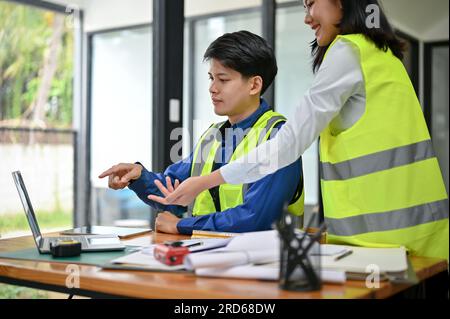 Un ingénieur asiatique intelligent et professionnel dans un gilet de sécurité discute d'un plan de construction sur un bureau avec sa collègue féminine au bureau. Banque D'Images