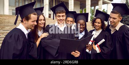 Heureux étudiants diplômés en robes noires de graduation cherchant à obtenir le diplôme de leur camarade de classe à l'extérieur. Banque D'Images