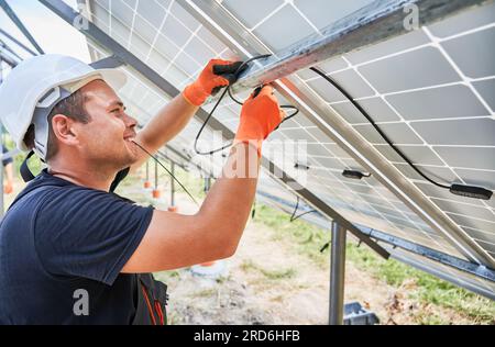 Close up of worker wiring solar panels together. Man installing photovoltaic solar panel system outdoors, wearing safety construction helmet and work gloves. Concept of renewable energy sources. Stock Photo