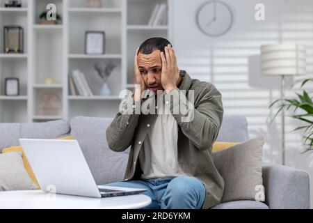 Jeune homme hispanique tenant sa tête choqué et inquiet, assis sur le canapé devant l'ordinateur portable à la maison et regardant l'écran bouleversé. Banque D'Images