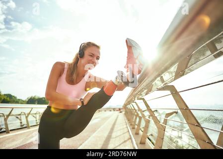 Femme handicapée positive en vêtements de sport et écouteurs étirant la jambe prothétique tout en se tenant debout sur le pont. Écouter de la musique Banque D'Images