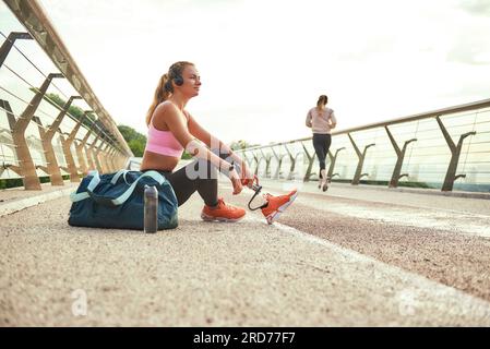 Il est temps de se détendre. Vue latérale de jeune belle femme dans des écouteurs avec prothèse de jambe écoutant de la musique et regardant de côté avec le sourire tout en étant assis sur le Banque D'Images