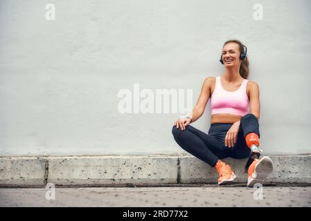 Simply Happy Jeune femme souriante avec prothèse de jambe dans des vêtements de sport et des écouteurs écoutant de la musique après son entraînement tout en étant assise à l'extérieur Banque D'Images