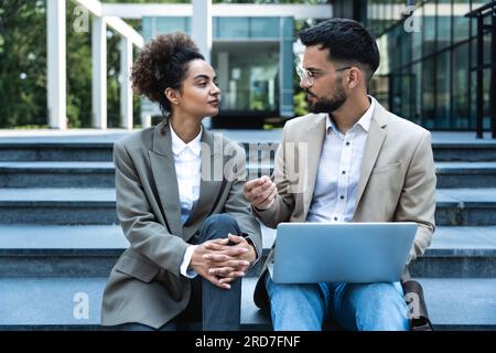 Bel homme et belle femme travaillant sur ordinateur portable sans fil près du centre de bureau. Deux collègues d'affaires assis dans les escaliers et travaillant à distance Banque D'Images