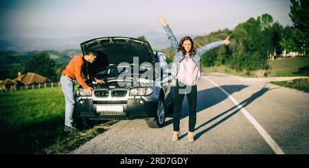 La voiture des jeunes couples est tombée en panne sur le chemin. Ils font de l'auto-stop pour trouver de l'aide. Le capot est ouvert et le moteur de la voiture est cassé Banque D'Images