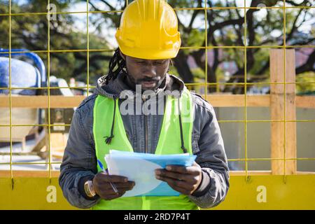 jeune ingénieur masculin de l'ethnicité africaine portant casque de sécurité jaune, veste et gilet, debout sur le chantier de construction avec dossier bleu repor de travail de lecture Banque D'Images