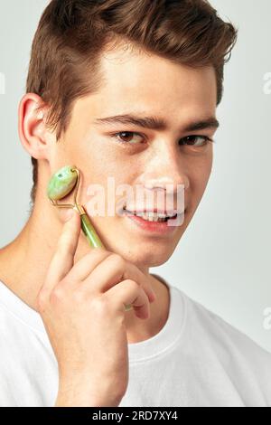 Jeune beau mec souriant aux cheveux bruns utilisant le rouleau de visage de dégonflement de jade dans le studio pour garder sa peau douce et lisse. Homme millénaire caucasien maintai Banque D'Images