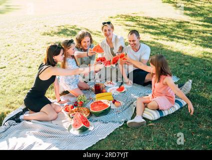 Les enfants avec la famille des parents couchés sur la couverture de pique-nique pendant le week-end journée ensoleillée, souriant, riant et levant des morceaux de pastèque juteux rouges Banque D'Images