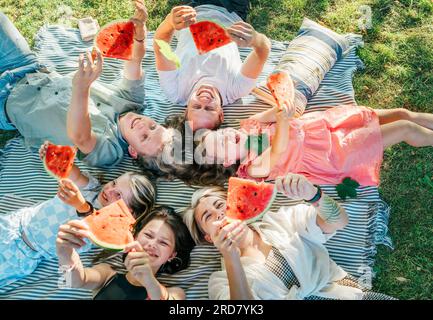 Les enfants avec la famille des parents couchés sur la couverture de pique-nique pendant le week-end journée ensoleillée, souriant, riant et levant des morceaux de pastèque juteux rouges Banque D'Images
