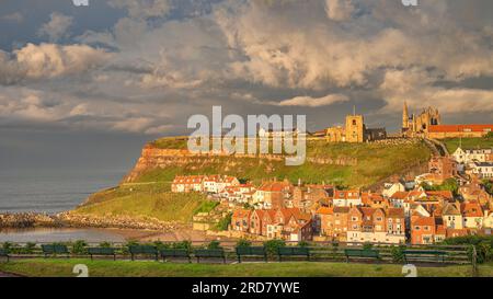 Le port extérieur de Whitby. Maisons nichées sous la falaise avec une église et l'abbaye de Whitby sur le sommet de la colline. Une ligne de bancs se trouve au premier plan. Banque D'Images