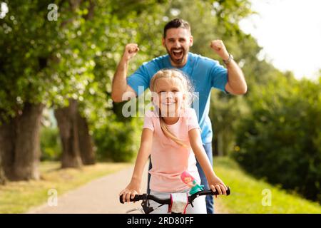 Heureux père enseignant fille à faire du vélo dans le parc, papa secouant les poings sur le fond, plaisir d'été et marcher à l'extérieur Banque D'Images