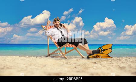 Homme d'affaires avec palmes de plongée et masque assis sur une chaise bech au bord de la mer faisant un appel téléphonique et pointant vers le haut Banque D'Images