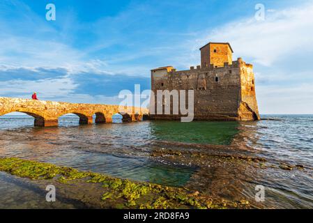 Homme sur le pont marche vers le château fort de Torre Astura dans l'eau de la mer Tyrrhénienne construite sur les ruines de villa romaine, heure du coucher du soleil Banque D'Images