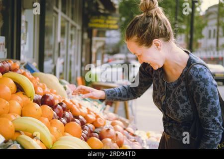 Heureuse jeune femme adulte riant en achetant des fruits dans un magasin d'alimentation, choisissant quoi acheter au stand de fruits sur un marché de rue, portant un sac fourre-tout. F Banque D'Images