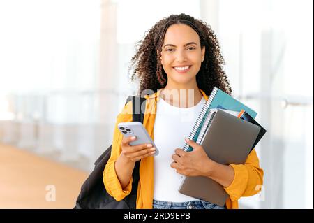 Heureuse magnifique étudiant de race mixte aux cheveux bouclés, avec un sac à dos, des livres et des cahiers dans sa main, debout près de l'université, tient smartphone, messagerie dans les médias sociaux, sourit à la caméra Banque D'Images