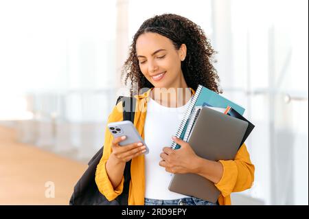 Positive jolie étudiante de race mixte aux cheveux bouclés, avec un sac à dos, des livres et des cahiers à la main, debout près du campus universitaire, tient un smartphone, messagerie dans les médias sociaux, sourit Banque D'Images