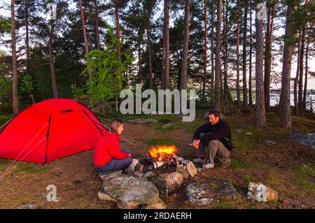 Un jeune couple campant dans la nature sauvage suédoise, assis autour d'un feu de joie crépitant ; profitant de la chaleur et de la tranquillité de leurs vacances d'été. Banque D'Images