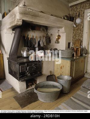 Vieille cuisine de l'époque avant les cuisinières électriques et les commodités modernes. Un cadre historique et une chambre avec une cuisine à l’ancienne où une cheminée en brique et un poêle à bois occupent le plus de place. Dans la chaleur du poêle, les chaussettes pendent pour sécher. Un pot de cuivre de bouillie est sur le poêle. Au sol, le lutefisk est en trempage dans l'eau dans une cuve en zinc. Un environnement qui était un spectacle commun du tournant du siècle 1800-1900 jusqu'aux années 1930 avec un mélange de différents objets de l'époque entre les deux. Banque D'Images