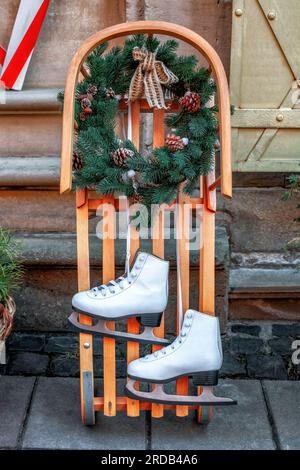 Patins à glace à deux chiffres, luge en bois, couronne de Noël. Paire de chaussures de skate en cuir blanc suspendues, traîneau en bois décorant avec couronne de sapin. Vintage hiver Banque D'Images