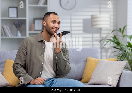 Jeune homme afro-américain assis sur un canapé à la maison et parlant sur un téléphone haut-parleur, enregistrant un message vocal, dictant sa conversation sur une application mobile. Banque D'Images