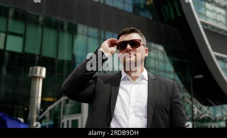 Homme d'affaires réussi dans un costume met des lunettes de soleil. Bel homme d'affaires mûr debout devant un gratte-ciel. Jeune homme d'affaires réussi met Banque D'Images