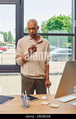 myasthénie gravis, homme d'affaires afro-américain audacieux avec le syndrome des yeux tenant smartphone et debout avec la canne de marche près du moniteur, la peau foncée de Banque D'Images