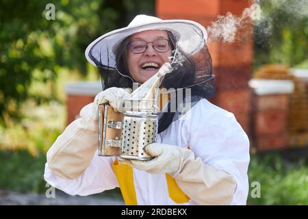 Une apicultrice âgée joyeuse tenant un fumoir dans le jardin de rucher en été Banque D'Images