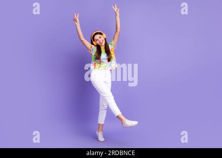 Photo pleine longueur de t-shirt imprimé fleur fille satisfait dans les écouteurs levant les mains vers le haut montrer v-sign isolé sur fond de couleur violette Banque D'Images