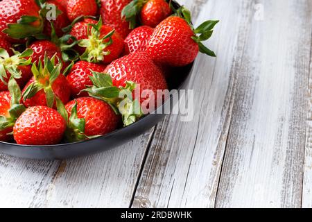 Grandes baies juteuses mûres de fraises biologiques dans une assiette ronde noire sur un fond en bois clair. Banque D'Images