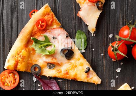 Les tranches de pizza au jerky, aux olives, au fromage et aux herbes sont disposées sur un fond en bois sombre, une branche de tomates cerises fraîches Banque D'Images