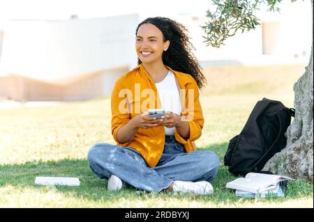 Étudiante ou freelance mixte aux cheveux bouclés positive et élégante, assise à l'extérieur sur l'herbe, utilisant son smartphone, messagerie sur les réseaux sociaux, SMS avec des amis, regarde loin, sourit joyeusement Banque D'Images