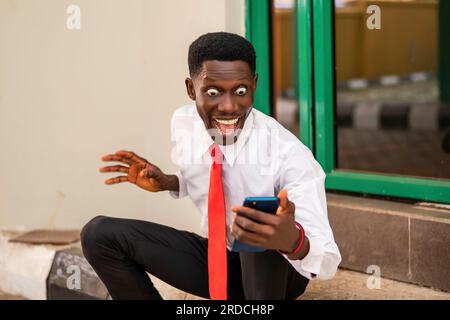 Photo d'un jeune homme africain qui semble excité tout en tenant un téléphone portable dans une chemise blanche et une cravate rouge Banque D'Images