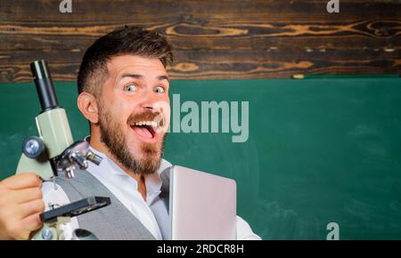 Professeur masculin heureux avec ordinateur portable et microscope. Étudiant avec cahier et microscope dans la salle de classe de l'école près du tableau avec espace de copie. Université Banque D'Images