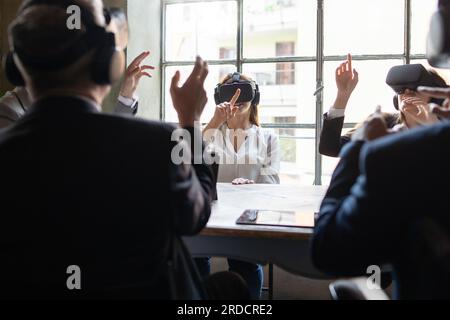 Équipe diversifiée de collègues dans une réunion d'affaires de réalité virtuelle dans un bureau. Tous portent des casques VR et font des gestes avec leurs mains. Banque D'Images