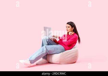 Jeune programmeuse féminine avec ordinateur portable assis sur pouf sur fond rose Banque D'Images