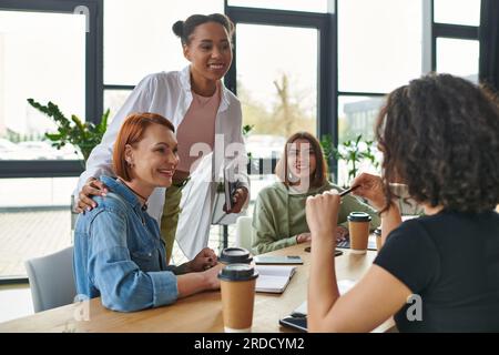 coach afro-américain joyeux avec carnet embrassant la femme et parlant à un groupe multiculturel assis près des boissons à emporter dans le club d'intérêt, mutuelles s Banque D'Images