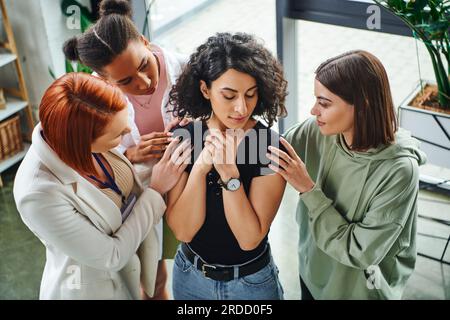 vue à grand angle du psychologue et des amies féminines multiethniques calmant la femme multiraciale frustrée debout les yeux fermés dans la salle de consultation, mora Banque D'Images